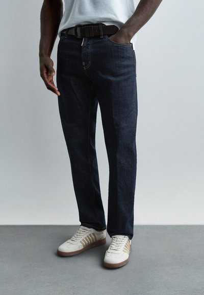Homme portant un jean bleu foncé, des baskets blanches avec des bandes marron, une ceinture noire et une chemise blanche, debout devant un fond gris uni.