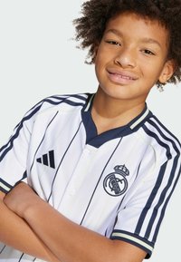 Maglia bianca Adidas con strisce navy, colletto a V, bottoni e logo ricamato del Real Madrid con dettaglio della corona sul lato sinistro del petto.