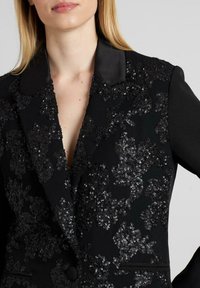 Blazer negro con cuello de satén, adornado con un patrón floral de lentejuelas negras. Presenta un cierre de botón único y detalles de bolsillos.