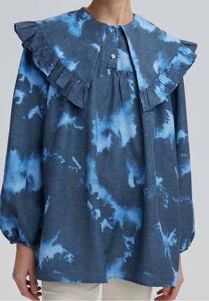 Niebieska bluzka tie-dye z dużym falbaniastym kołnierzem, długimi bufiaste rękawami, zapinanym dekoltem i elastycznymi mankietami, noszona na jasne spodnie.