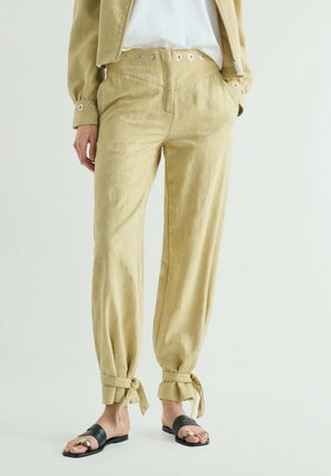 Pantalones beige de tiro alto con detalles de ojales, lazos en los tobillos, combinados con una chaqueta a juego, top blanco y sandalias negras abiertas en los dedos.