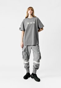Camiseta gris de gran tamaño con el texto "MWM", combinada con pantalones cargo grises que cuentan con múltiples bolsillos y acentos en blanco, y calzado negro.