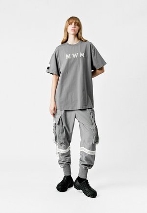 Camiseta gris de gran tamaño con el texto "MWM", combinada con pantalones cargo grises que cuentan con múltiples bolsillos y acentos en blanco, y calzado negro.