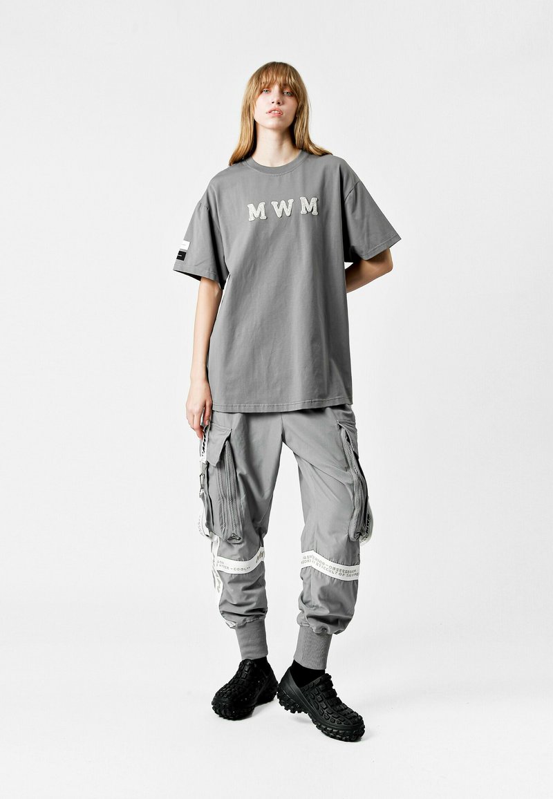 Camiseta gris de gran tamaño con el texto "MWM", combinada con pantalones cargo grises que cuentan con múltiples bolsillos y acentos en blanco, y calzado negro.