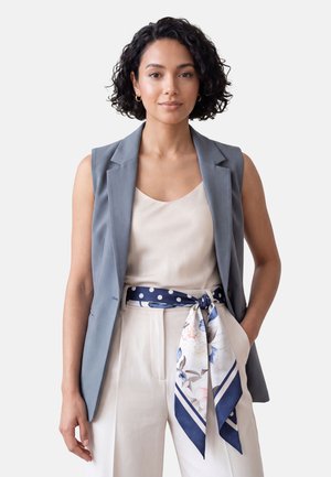 Mujer con pelo rizado que lleva una camiseta beige claro, un chaleco gris sin mangas, pantalones blancos y un cinturón de bufanda azul con flores, de pie con una mano en el bolsillo.