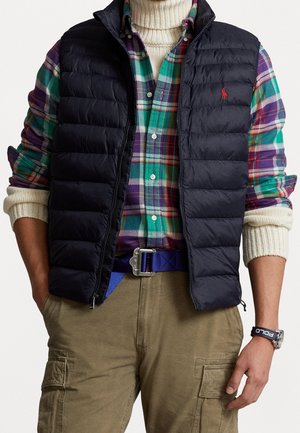 Mann, der geschichtete Kleidung trägt: cremefarbener Rollkragenpullover, grün-lila kariertes Hemd, schwarze gesteppte Weste, beigefarbene Cargohose und blauer Gürtel.