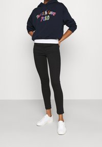 Marinblå hoodie med grafiken "Peace Love Polo" i flerfärg, kombinerad med svarta skinny jeans och vita sneakers.