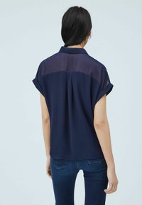 Navy blue short-sleeve shirt with a sheer back panel, collar, and rolled cuffs. Smooth texture with a loose fit, paired with denim jeans.