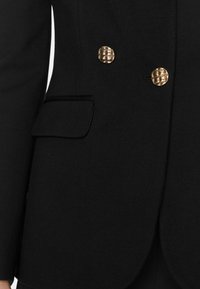 Blazer nero su misura dal design elegante, con due bottoni rotondi dorati e una piccola tasca a pattina sul lato sinistro.