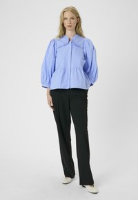 Blusa a righe azzurro chiaro con maniche a sbuffo, colletto arricciato e vita peplo, abbinata a pantaloni neri sartoriali e scarpe basse bianche con punta.