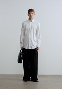 Άσπρο πουκάμισο με μακριά μανίκια και κουμπιά και μαύρο παντελόνι με φαρδιά πόδια, κρατώντας μια μαύρη δερμάτινη τσάντα με κυκλικό χερούλι, σε απλο φόντο.