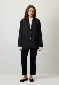 Melna žakete ar zeltītām pogām, apvienota ar krēmkrāsas ribotu sweateri, melnu korduroja biksēm un brūnām leopardu raksta čībām. Vienkāršs fons.