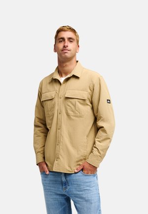 Homme aux cheveux courts châtain clair portant une veste beige boutonnée avec poches poitrine et un jean bleu, debout devant un fond clair uni.