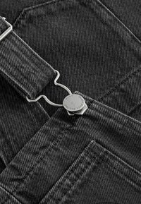 Schwarze Denim-Overallträger mit einem silbernen Metallclip und Beschlägen. Der Stoff hat ein strukturiertes, strapazierfähiges Aussehen mit sichtbaren Nähten.