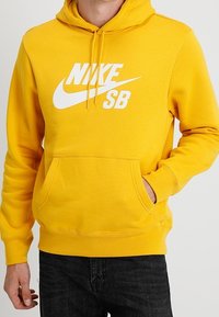 Man i gul Nike SB-hoodie med framficka och svarta jeans, hand delvis i fickan, mot en enkel bakgrund.
