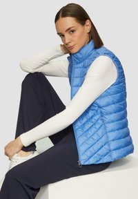 Gilet bouffant bleu clair avec des coutures verticales, col haut et poches latérales zippées, porté sur un haut à manches longues blanches à côtes.