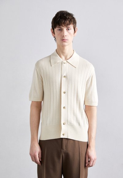 Polo din tricot cu mânecă scurtă, de culoare crem, cu caneluri verticale, cinci nasturi aurii și guler punctat. Fabricat dintr-un material moale și texturat.