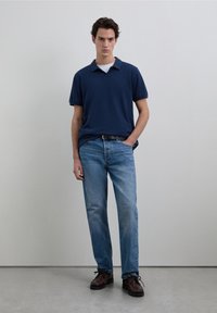 Polo azul marino de manga corta sobre una camiseta blanca, combinado con jeans rectos de color azul claro y zapatos marrones con cordones.