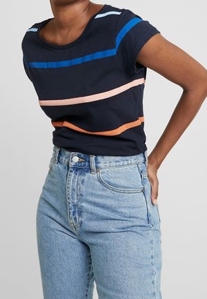 Person trägt ein dunkelblaues T-Shirt mit horizontalen blauen, pinken und orangenen Streifen kombiniert mit hoch taillierten hellblauen Jeans, Hand ruht am Rücken.
