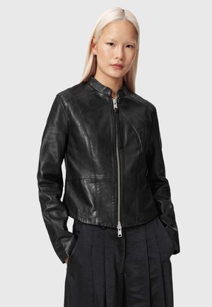 Vrouw met lang blond haar, gekleed in een nauwsluitend zwart leren jack met een rits aan de voorkant en donkere geplooide broek, staand met handen in de zakken.