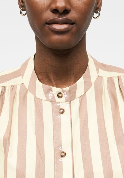 Κοντινό πλάνο ενός ατόμου που φοράει χρυσούς κρίκους για σκουλαρίκια και πουκάμισο με κάθετες ρίγες σε μπεζ και κρεμ χρώμα με στρογγυλά καφέ κουμπιά.