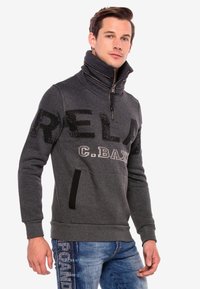 Grauer Pullover mit hohem Kragen, Reißverschluss und schwarzem Pailletten-Schriftzug. Verfügt über Seitentaschen sowie gerippte Bündchen und Saum.
