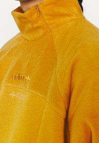 Keltainen fleecepulloveri, jossa korkea kaulus, vetoketjukiinnitys ja kirjailutettuja logoja. Pehmeä rakenne, urheilullinen muotoilu ja sauma-yksityiskohdat.