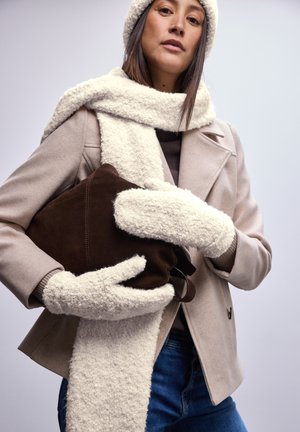 Cremefarvet tørklæde og handsker lavet af blødt materiale. Personen bærer en lys beige frakke og holder en mørkebrun taske. Blå jeans er synlige.