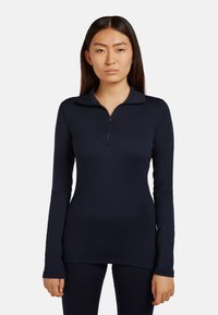 Jeune femme aux cheveux lisses et foncés portant un haut bleu marine à manches longues ajusté avec fermeture éclair et un pantalon assorti, posant devant un fond uni.