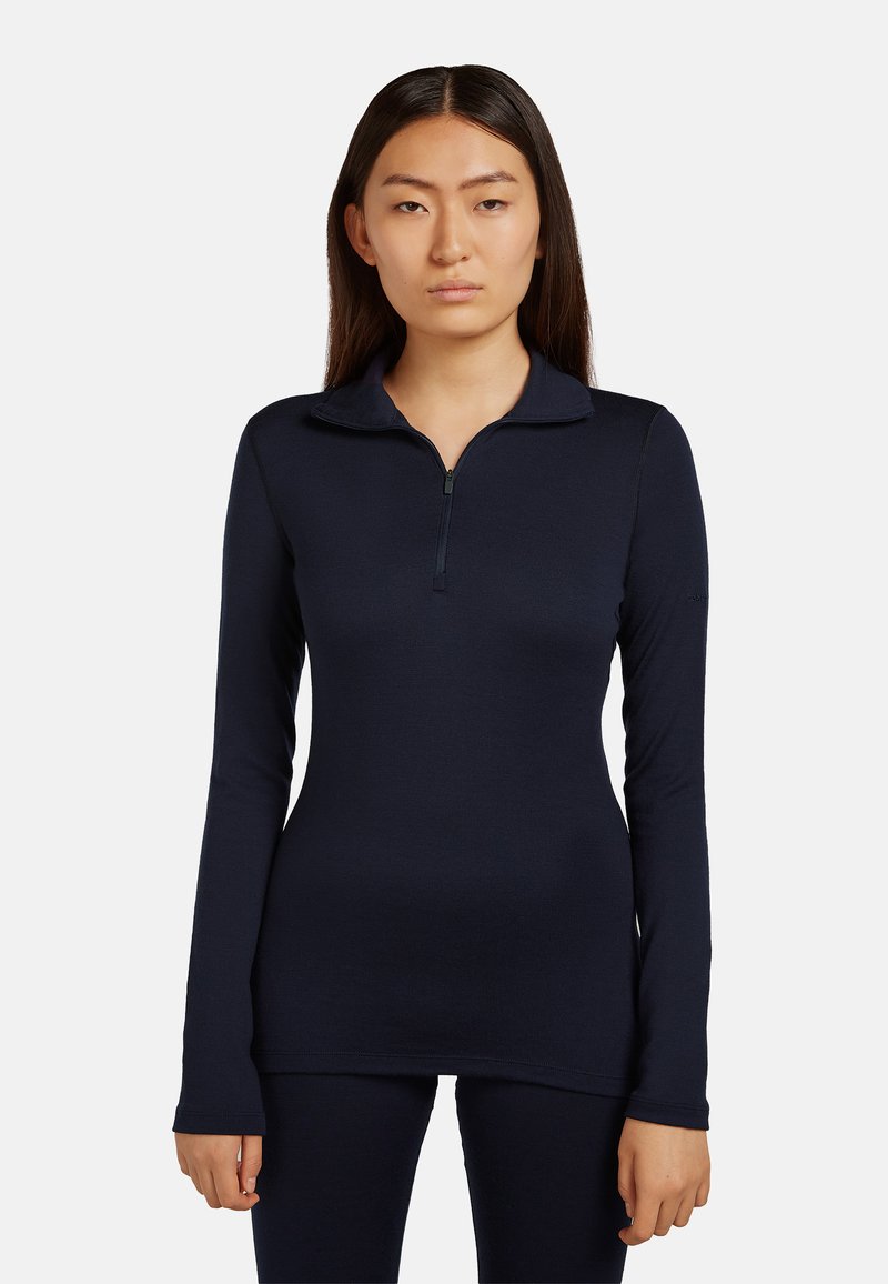 Jeune femme aux cheveux lisses et foncés portant un haut bleu marine à manches longues ajusté avec fermeture éclair et un pantalon assorti, posant devant un fond uni.