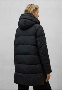 Osoba v černé prošívané bundě s kapucí, otočená zády, s krátkými blond vlasy, na jednobarevném světle šedém pozadí.