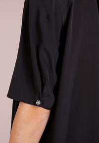 Camisa negra de manga corta, con un puño fruncido adornado con un pequeño acento decorativo de strass. Tela de textura suave.
