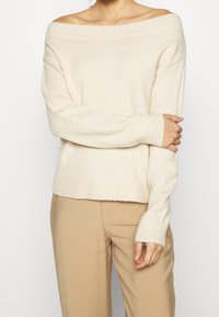 Krämfärgad off-shoulder-tröja med ribbade ärmslut och nederkant, med mjuk textur och avslappnad passform, i kombination med beigefärgade byxor.