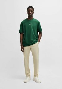 Grønn t-skjorte med korte ermer med "BOSS"-logo, kombinert med lysebeige bukser og hvite sneakers. Enkel design; glatt stoffstruktur.
