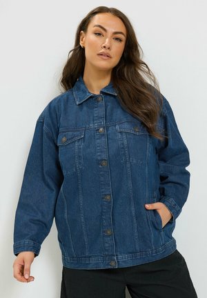 Femme aux longs cheveux bruns portant une veste en jean bleu foncé avec des boutons et des poches sur le devant, debout devant un fond uni clair.