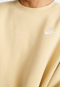 Muž v béžovém svetru s kulatým výstřihem, na levé části hrudi bílá logo Nike, detailní záběr horní části trupu a výstřihu.