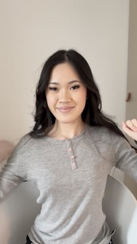 Jeune femme aux longs cheveux foncés sourit et ajuste sa chemise grise à manches longues tout en étant assise contre un mur beige uni.