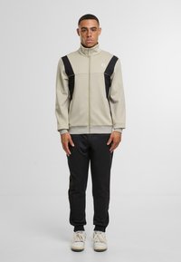 Pánský teplákový overal se béžovou bundou na zip s �černými akcenty a odpovídajícími černými kalhotami. Materiál má hladkou texturu a přiléhavý střih.