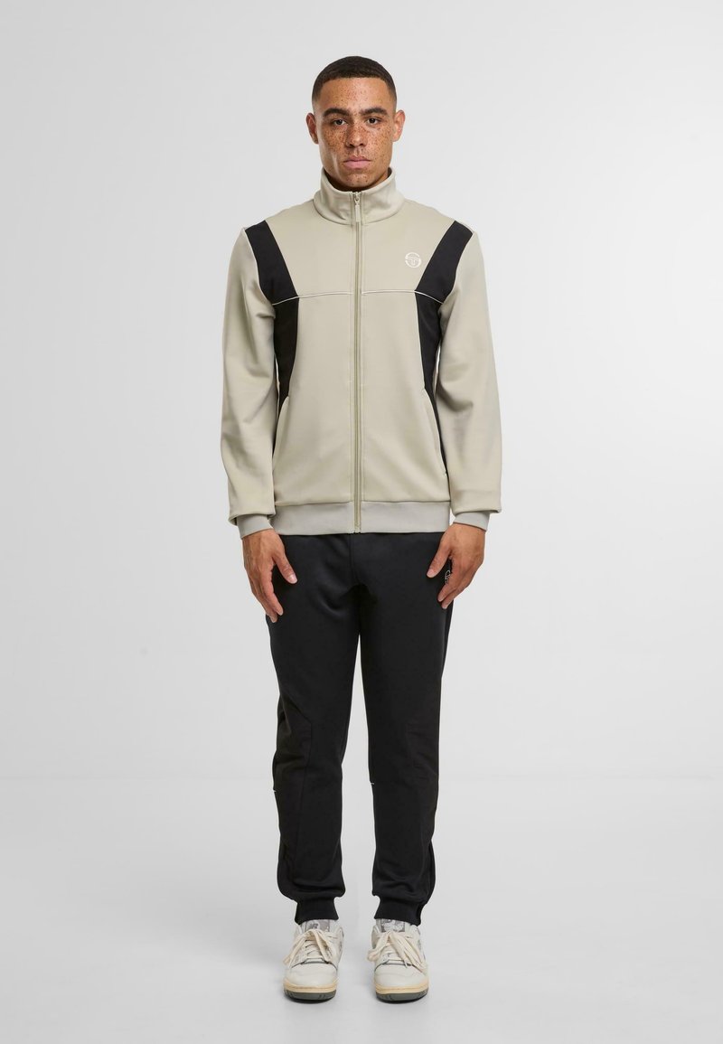 Pánský teplákový overal se béžovou bundou na zip s černými akcenty a odpovídajícími černými kalhotami. Materiál má hladkou texturu a přiléhavý střih.