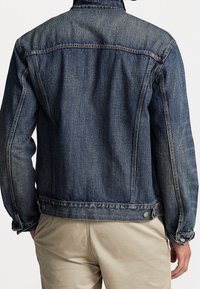 Muž oblečený v modré džínové bundě a béžových kalhotách, zobrazený zezadu na jednoduchém bílém pozadí.