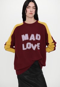Burgundy knitted sweater with yellow ribbed sleeves, featuring the text "MAD LOVE" in distressed light purple. Styled with a black skirt.