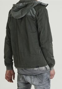 Ciemnozielona kurtka z kapturem o teksturowanym wykończeniu, z elastycznymi mankietami i zaokrąglonym dołem. Noszona na paskowanej koszuli i szarych dżinsach.