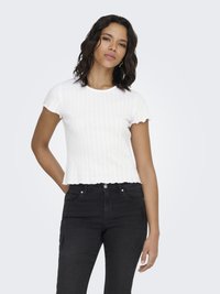 Vrouw met golvend donker haar, gekleed in een wit geribd topje met korte mouwen en een zwarte hoge taille broek, staand met �één hand achter haar rug.