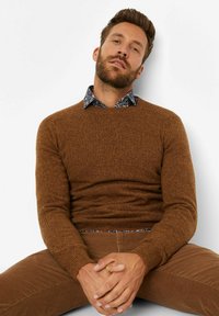 Brauner Strickpullover mit gerippten Bündchen, über einem blauen blumigen Hemd mit Kragen getragen. Model sitzt, die Hände gefaltet, in Cordhosen.