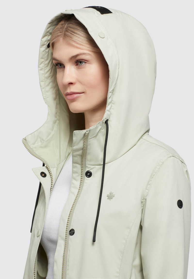 Giacca verde chiaro con cappuccio, frontale con zip, bottoni a pressione e cordini neri; presenta un piccolo logo sul petto e dettagli neri.