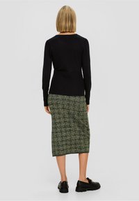 Kobieta z blond bobem, ubrana w czarną bluzkę z długim rękawem, zieloną spódnicę midi w wzory i czarne loafersy, stojąca na tle jednolitym.