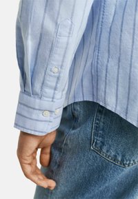 Camicia azzurra con bottoni, a righe bianche verticali, con polsini a punta e bottoni doppi, abbinata a jeans in denim blu.