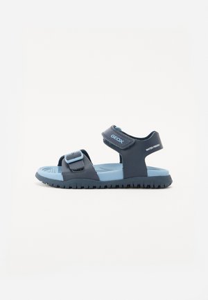 Sandal de cor azul escuro com tiras ajustáveis, sola de borracha e palmilha acolchoada. Apresenta a marca "GEOX" e elementos de design à prova de água.