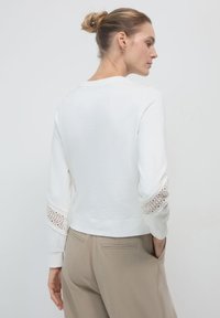 Donna con un chignon biondo, indossa una maglia bianca a maniche lunghe con dettagli di pizzo sulle maniche e pantaloni beige, in piedi con una mano in tasca.