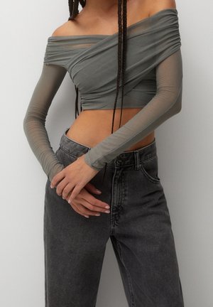 Kvinde iført en gennemsigtig, langærmet grå off-shoulder crop top og højttaljede mørkegrå jeans med hænderne foldet foran.